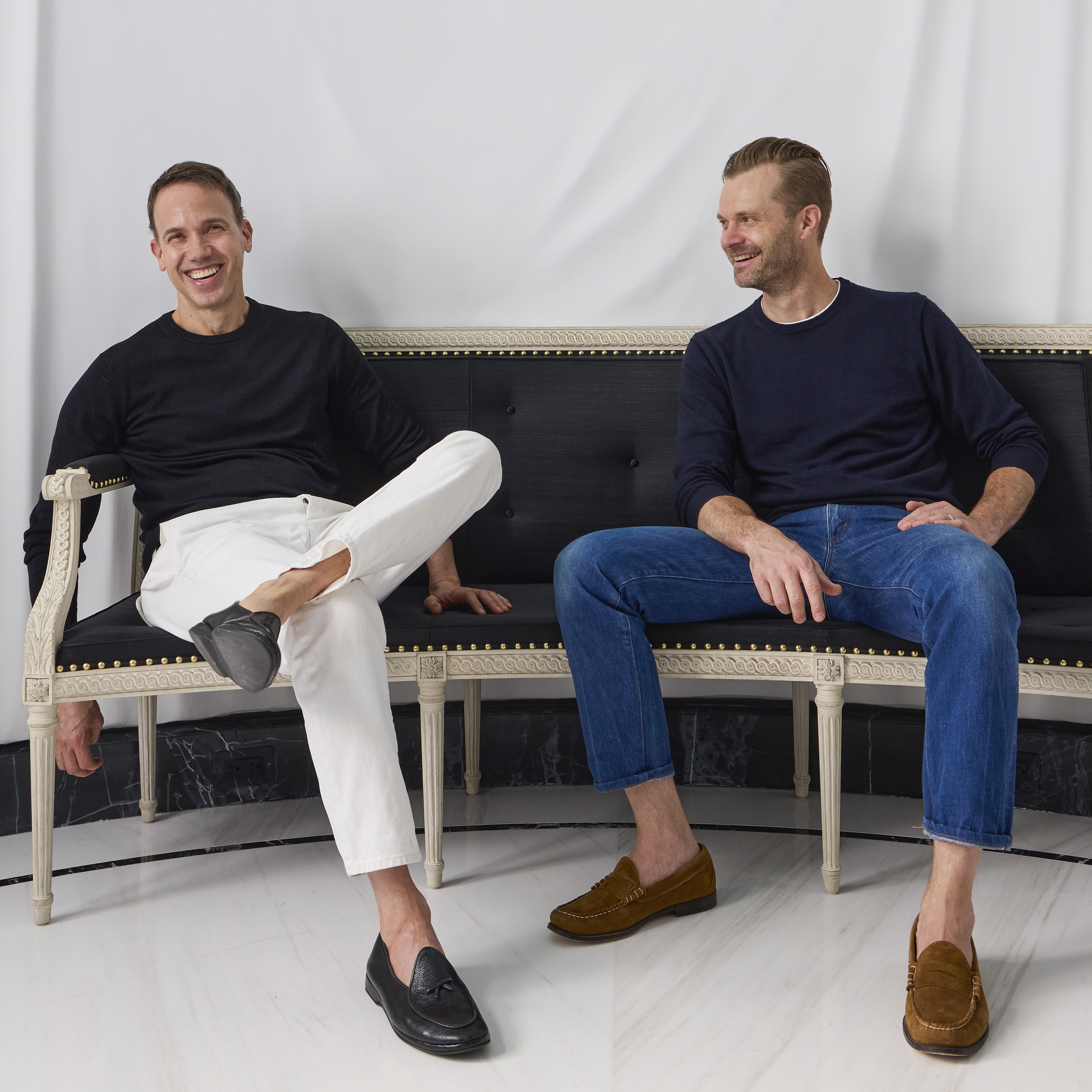 two men sitting on a bench in front of a white backdrop